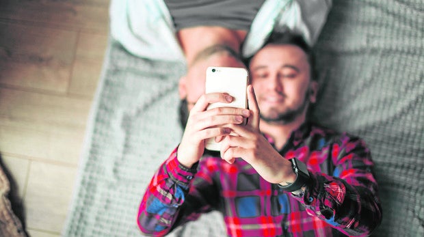 Un hombre tumbado en la cama, junto a otro, mira su móvil