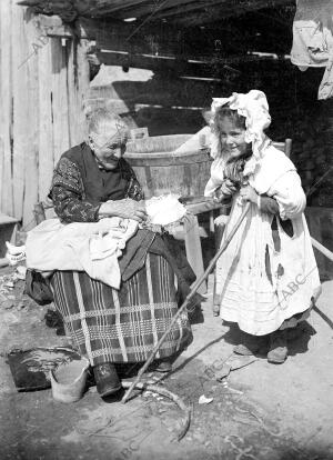 En la imagen, una anciana con una muñeca y una niña con un báculo y una cofia...