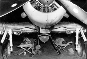 Mecánicos norteamericanos descansan bajo su avión, con fusiles y cartucheras a...