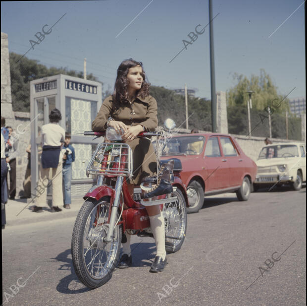 Una joven en una vespino, con unos patines para hielo colgados del hombro, con...